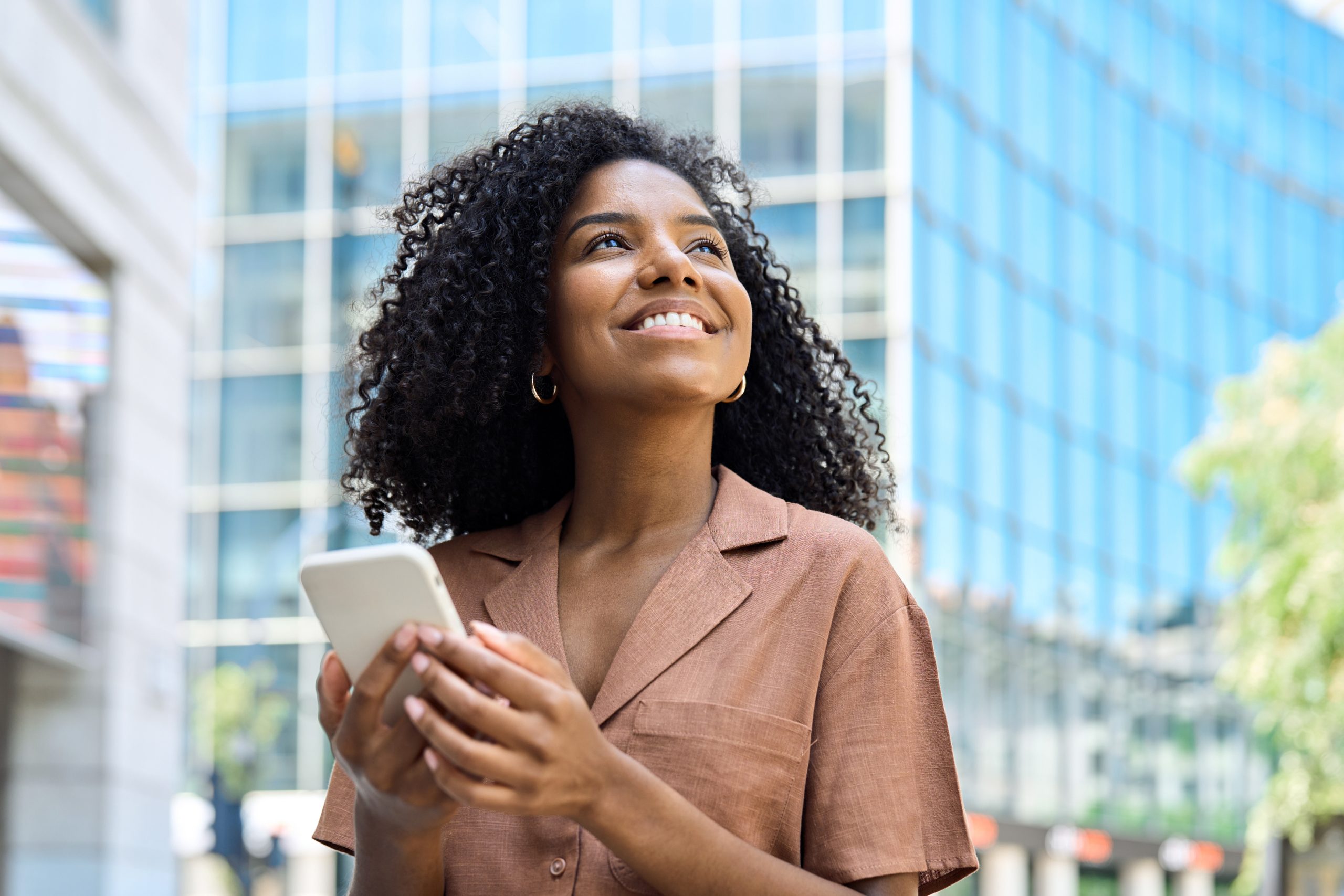 Happy young African woman using smartphone standing in city looking away.