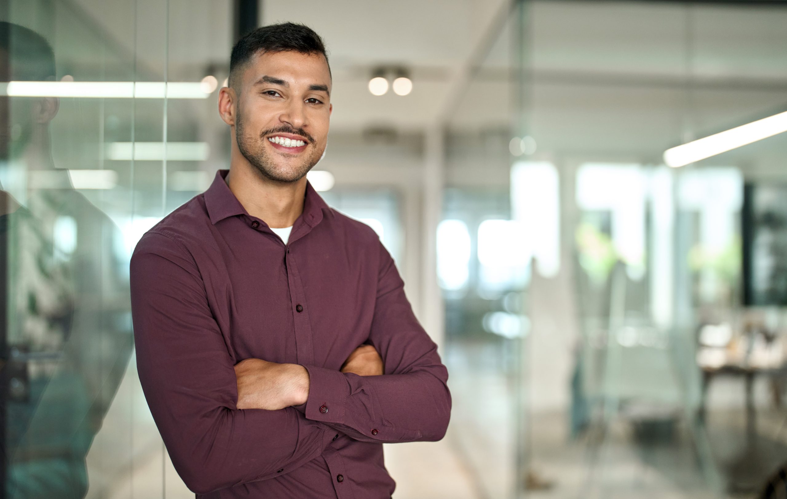 Confident young Latin business man standing arms crossed in office, portrait.