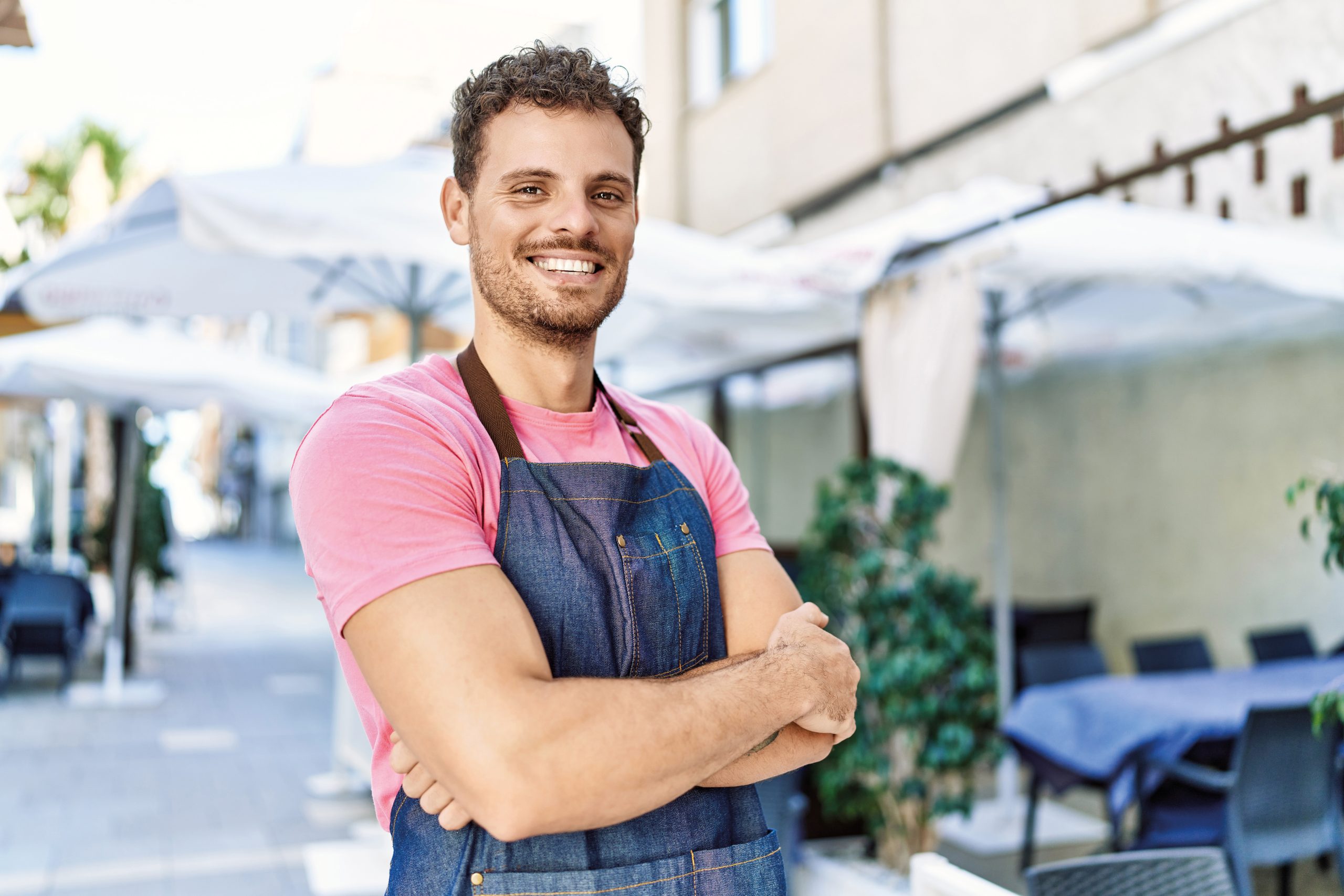 Young hispanic waiter wearing apron standing with arms crossed gesture at coffee shop.